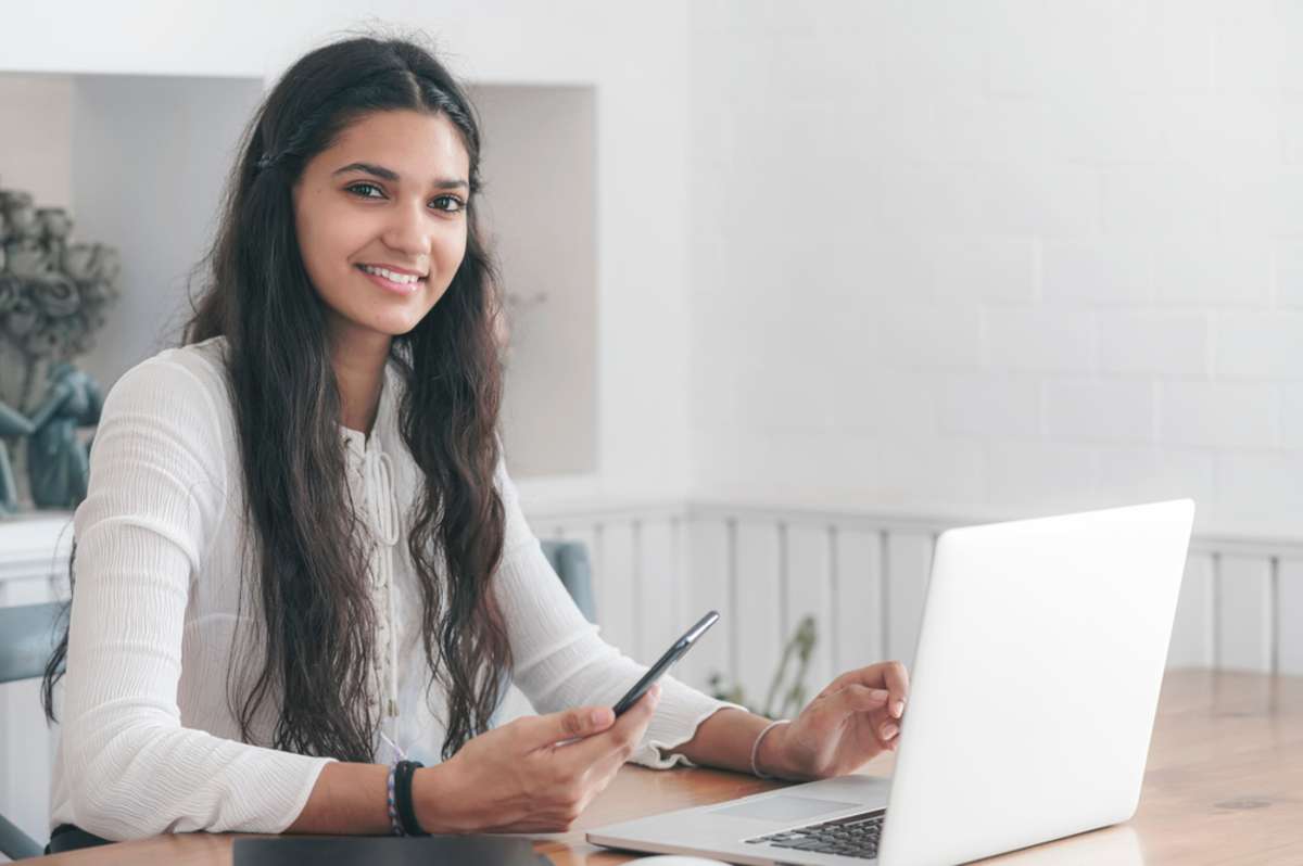 Attractive smiling young african woman working on laptop computer and using smartphone at the table at home