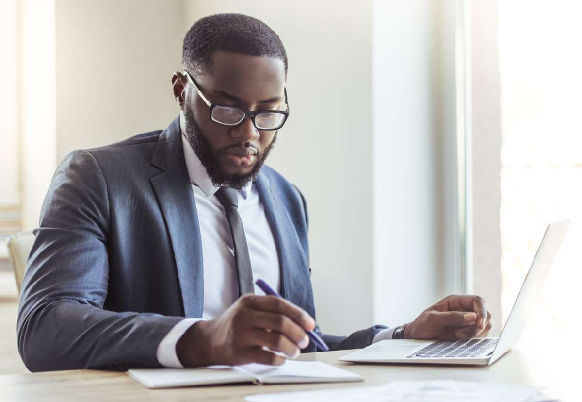 Handsome Afro American businessman in classic suit and eyeglasses is using a laptop and making notes while working in office