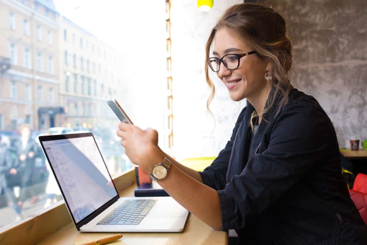 Happy smiling woman in glasses skilled business worker checking e-mail on mobile phone while sitting with laptop computer in coffee shop interior