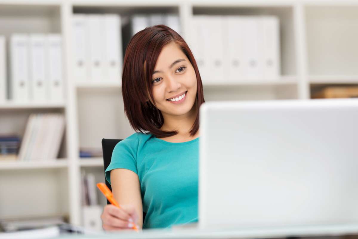 Smiling relaxed young Asian businesswoman at work leaning back comfortably in her chair as she works at her laptop smiling in confidence